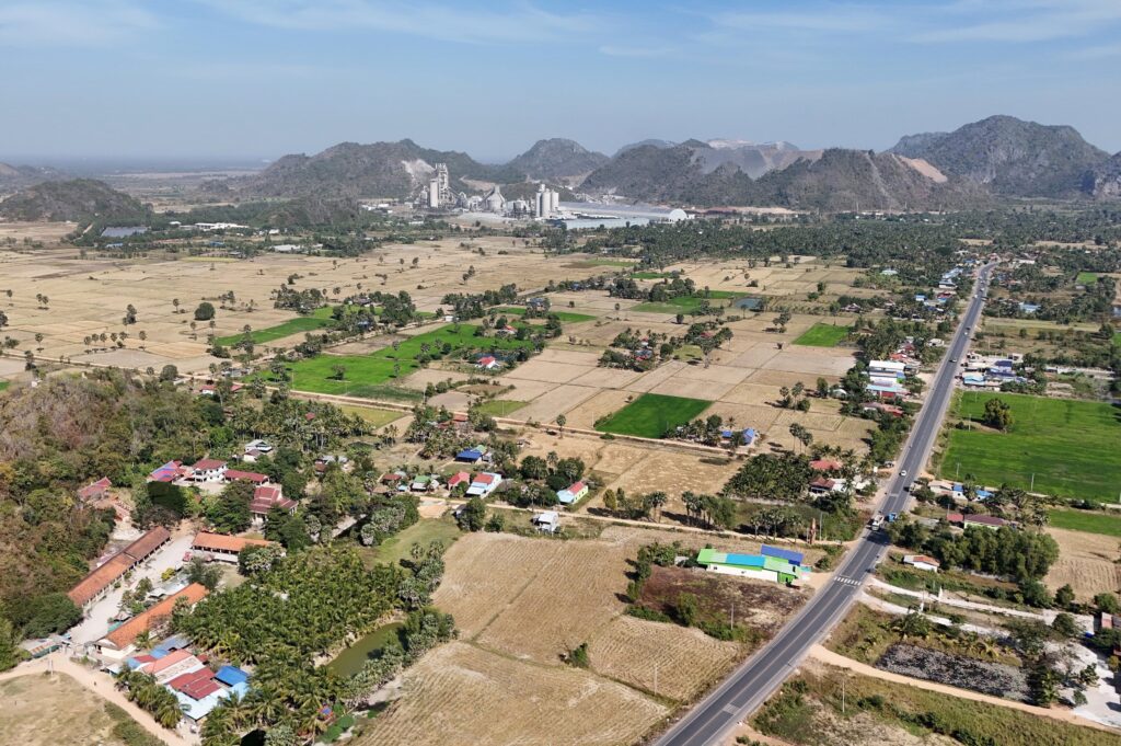 A tarmac road runs towards the cement kiln in the background, surrounded by houses and farmland.